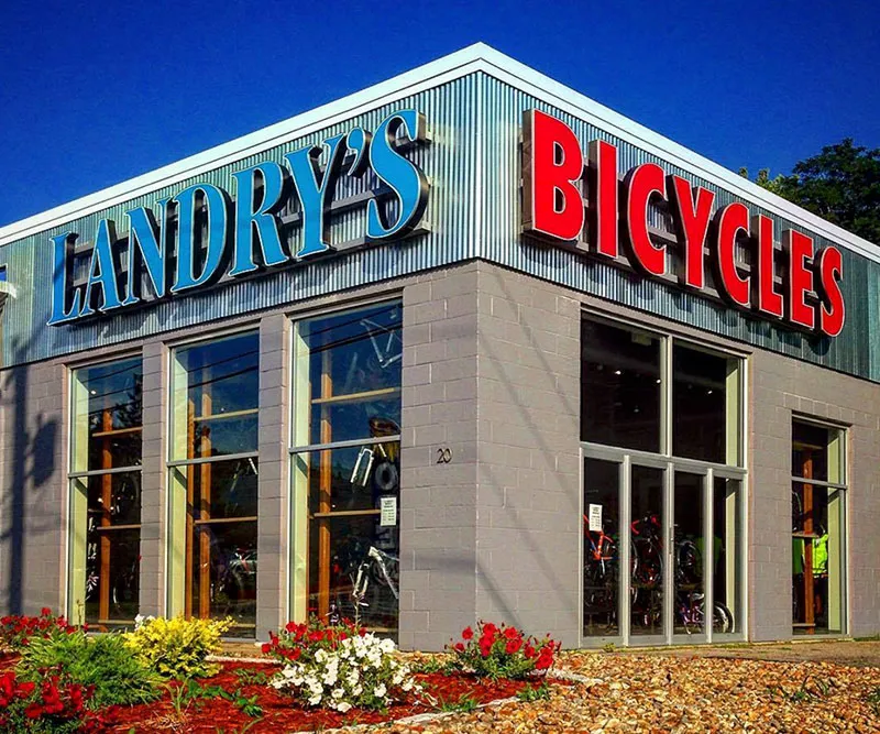 Landry's Bicycles storefront with blue and red signage, glass windows showing bikes, and flower bed in front.