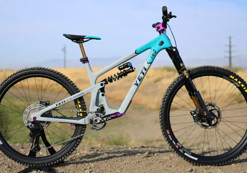 Side view of a gray and teal Yeti full-suspension mountain bike on a dirt trail with hills behind.