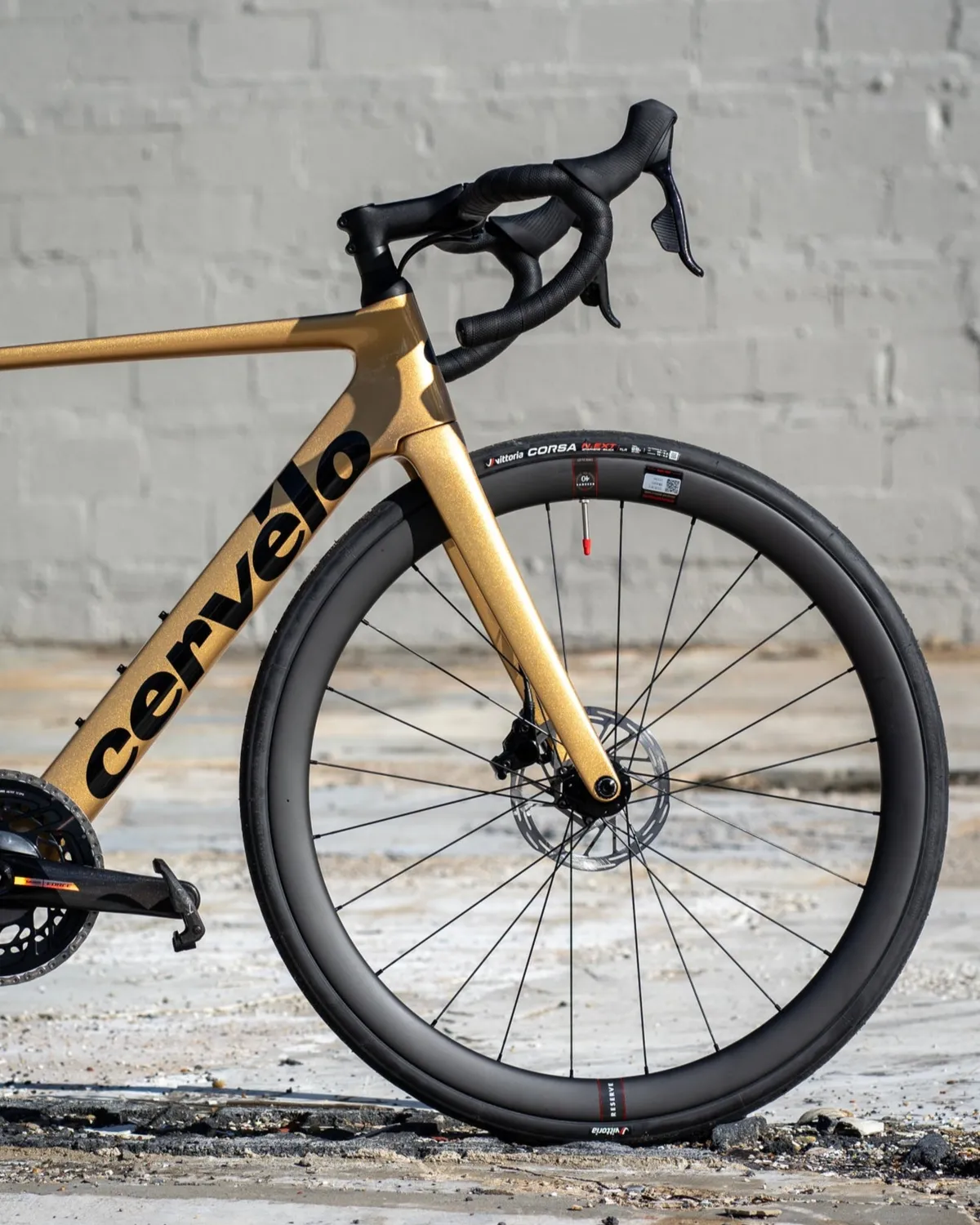 Metallic gold Cervélo road bike with black carbon wheel and drop handlebars against a grey brick wall.