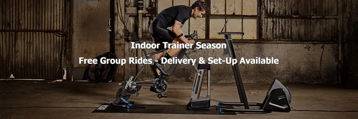 Cyclist on road bike using Wahoo KICKR trainer, fan, and desk in rustic room with promotional text overlay