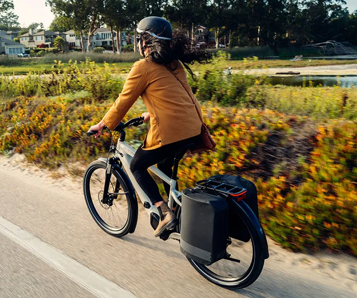 Woman in yellow jacket and helmet riding a white Specialized e-bike with rear panniers on a paved coastal path.